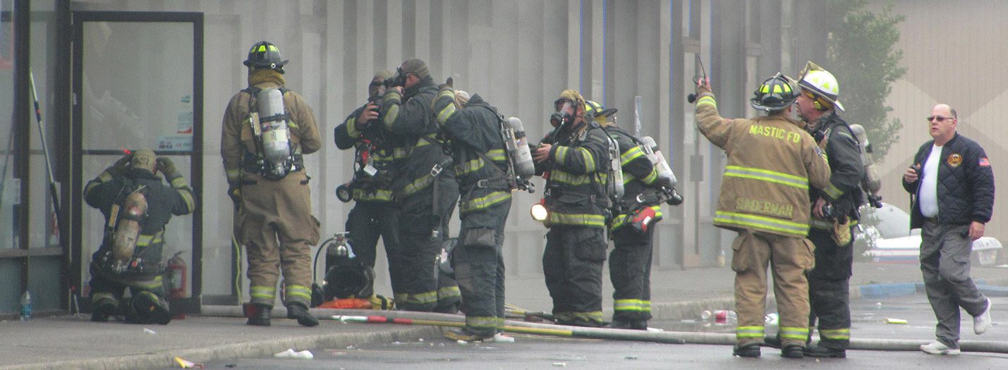 Several volunteer firefighters from Mastic FD outside a commercial building with smoke in the air