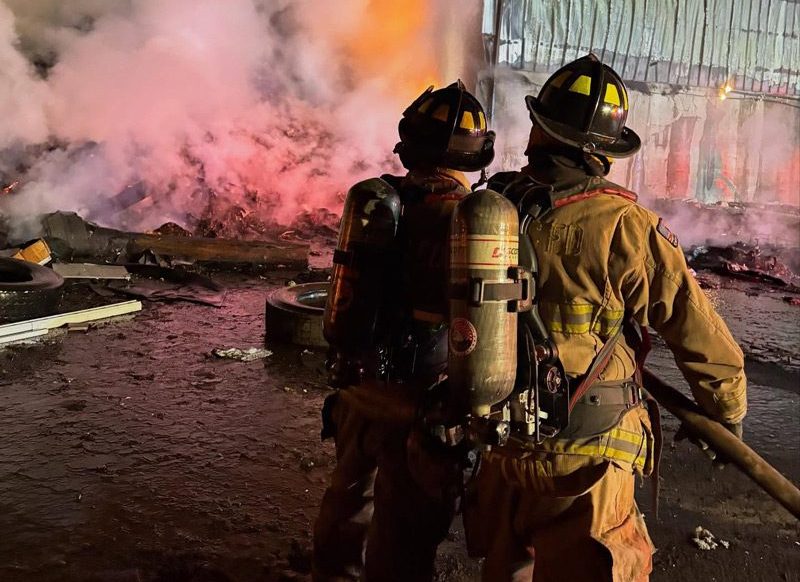 photo from behind 2 firefighters using a firehose to put out a fire